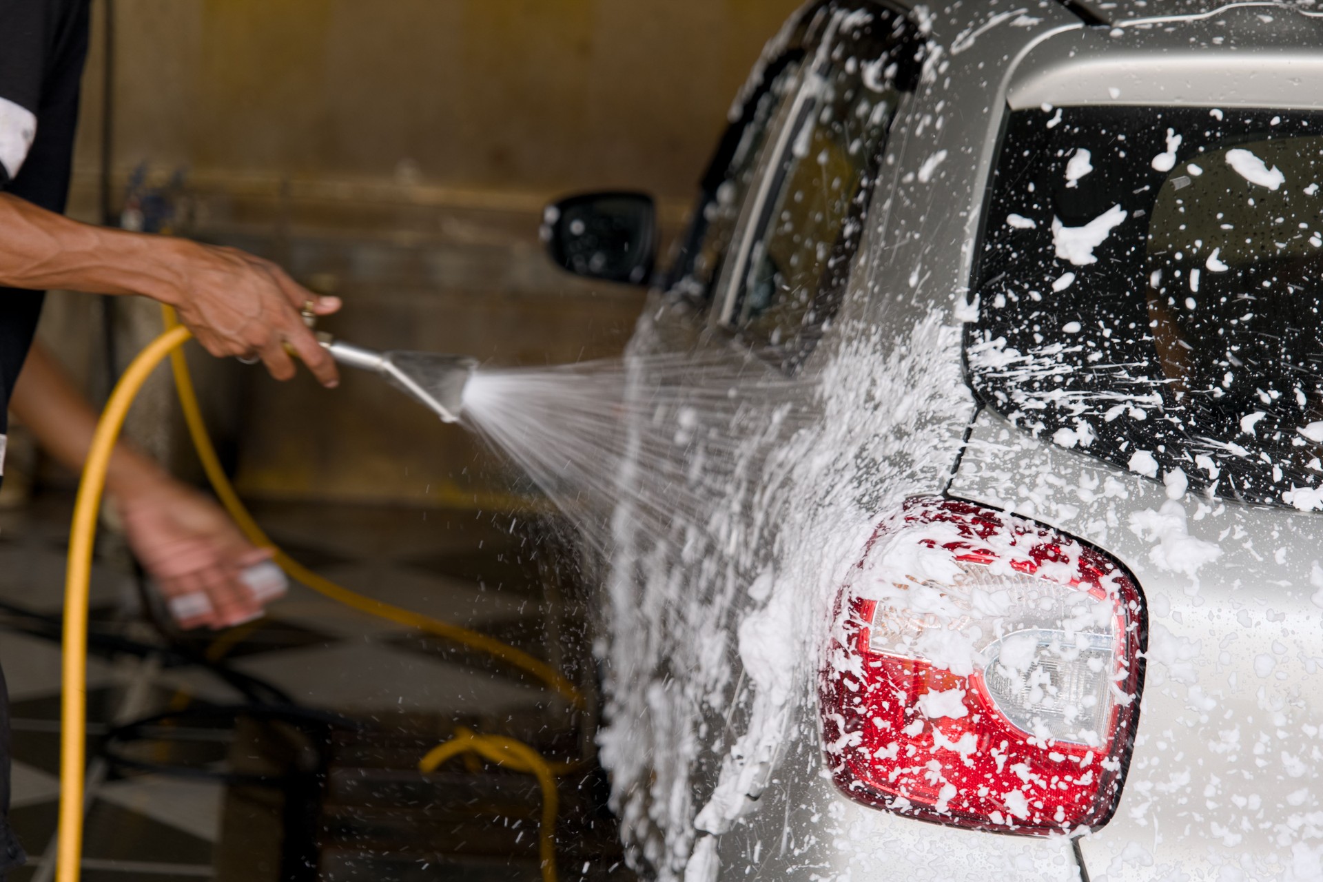 A person washing a silver car with a spray hose at a car wash facility during the day for thorough cleaning and maintenance